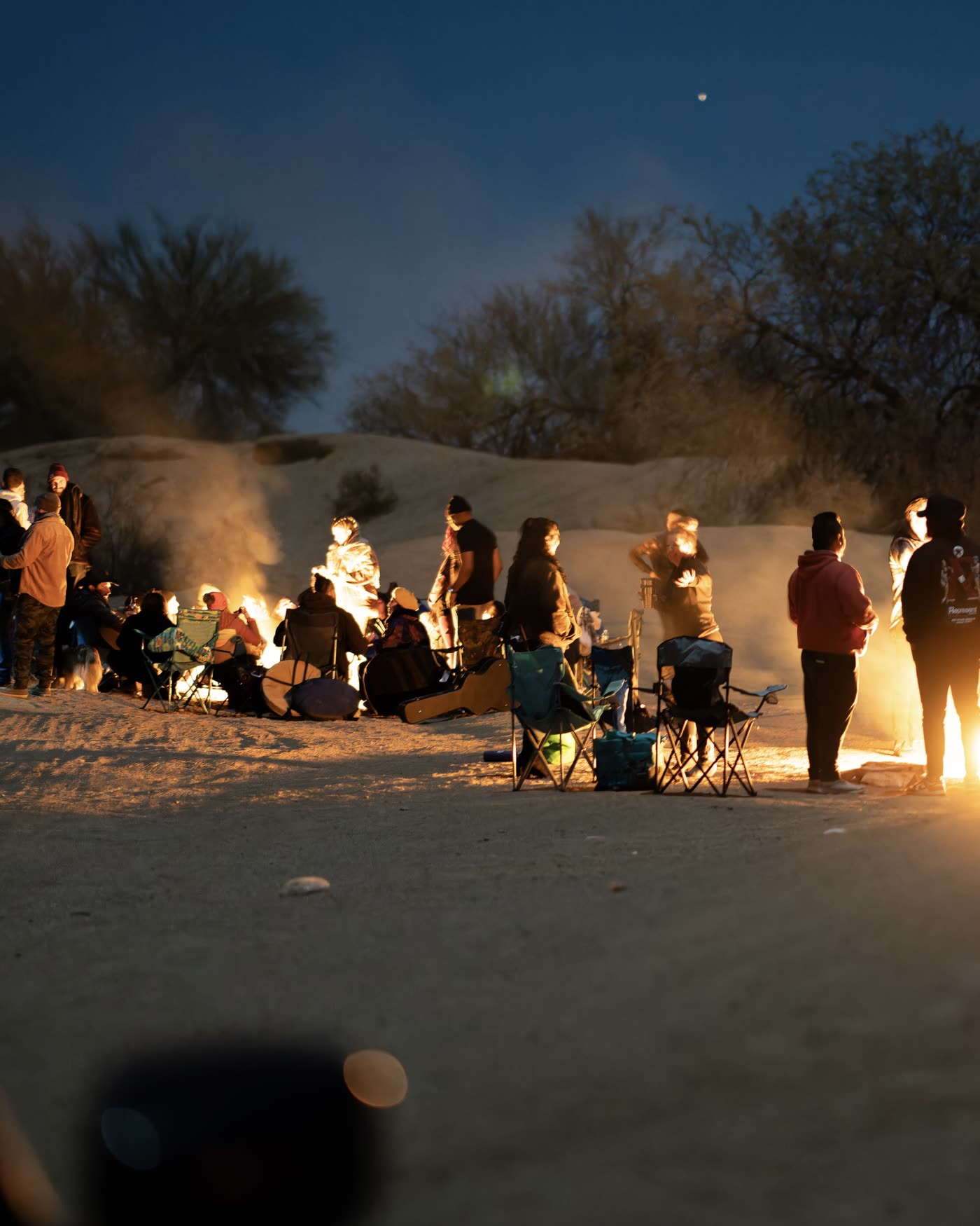 A crowd gathered around a fire at dusk in the open desert, moon overhead